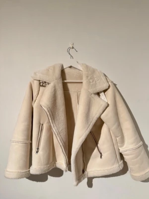 Beige teddyjacka med dragkedjor - Säljer en beige teddyjacka med fluffig insida och bred krage. Jackan har två snedställda dragkedjor fram och detaljer med spännen vid kragen. Materialet är mjukt och ger en cozy vibe, perfekt för kyliga dagar. Jackan har en loose passform och långärmade ärmar.