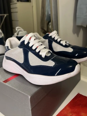 Prada sneakers - Säljer ett par snygga Prada sneakers i marinblått och vitt med röda detaljer. Skorna har en rund tå, platt sula och snörning. Ovandelen är i mesh och lackad syntet, vilket ger en modern och sportig vibe. Perfekt för dig som gillar exklusiva märken och stilren design.