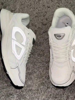 Vita Dior B30 sneakers - Säljer ett par vita Dior B30 sneakers med reflekterande detaljer och tydlig Dior-logga på sidan. Skorna har en chunky sula, meshpaneler och snygga kontrastsömmar. Perfekta för dig som vill ha en clean och exklusiv look.