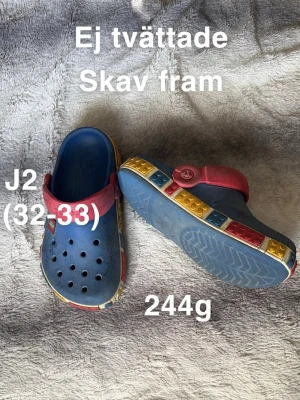 Blå Crocs med LEGO-detaljer - Säljer ett par blå Crocs med coola LEGO-inspirerade detaljer runt sulan i rött, gult och blått. Skorna har röd hälrem och ventilerande hål på ovansidan. Perfekt för dig som gillar färgglada och lekfulla skor.🌸 Använt skick. Hela. Skav fram. Ej tvättade storlek 32-33. Innermått ca 21.5cm 