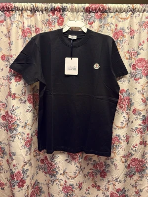 Svart t-shirt från Moncler XXL - Säljer en svart t-shirt från Moncler i storlek XXL. T-shirten har rund hals, korta ärmar och en clean look med Moncler-logga på bröstet. Materialet är mjuk bomull som känns skönt mot huden. Perfekt för dig som gillar stilrena och klassiska plagg.