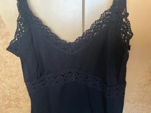 Svart linnetopp med spets från Vero Moda - Säljer en svart linnetopp från Vero Moda i storlek M. Toppen har breda axelband och v-ringad hals, båda dekorerade med snygg spets. Spetsdetaljer finns även runt midjan för extra stilpoäng. Perfekt att matcha med jeans eller kjol.