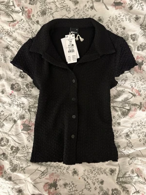 Svart kortärmad blus från Gina Tricot - Säljer en svart kortärmad blus från Gina Tricot med krage och knappar framtill. Toppen har en strukturerad, lite ribbad känsla och är i ett mjukt material som sitter snyggt på kroppen. Perfekt till jeans eller kjol för en enkel men stilren look.