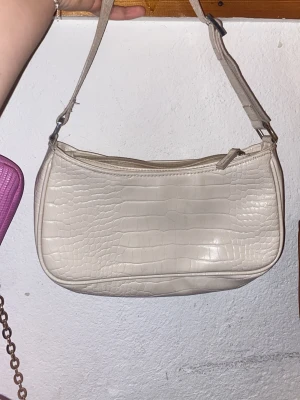 Beige axelväska med krokopräglat mönster - Säljer en beige axelväska med krokopräglat mönster och justerbar rem. Väskan har en stilren form och stängs med dragkedja upptill. Perfekt för att lyfta din outfit med en trendig touch. Materialet är syntet och ger en lyxig känsla.