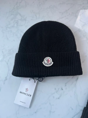 Svart ribbad mössa från Moncler - Svart ribbstickad mössa från Moncler med uppvikt kant och klassisk Moncler-logga framtill. Mössan är tillverkad i mjukt material och har en stilren look som passar perfekt till kalla dagar. Taggar och etikett medföljer.