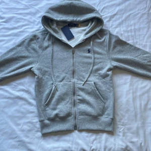 Ralph Lauren zipup hoodie grå - Snygg grå hoodie från Polo Ralph Lauren med dragkedja, huva och snörning. Tröjan har broderad blå logga på bröstet och två fickor framtill. Mjuk insida och klassisk passform, perfekt för chill dagar.