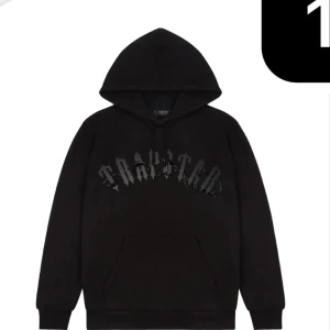 Svart Trapstar hoodie med tryck - Svart hoodie från Trapstar med ikoniskt tryck i grått framtill. Klassisk känguruficka, huva med snörning och mjukt material. Perfekt för dig som gillar streetwear och vill sticka ut med en edgy look.