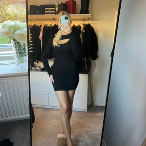 Svart ribbad kortklänning med cut-out - Snygg svart ribbad kortklänning med lång ärm och cool cut-out detalj vid halsen. Klänningen har en tight passform som framhäver figuren och är perfekt för dig som gillar minimalistisk stil med en twist.