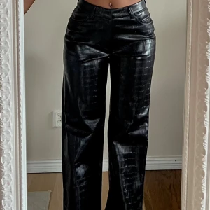 Svarta croc-mönstrade byxor i skinnimitation - Säljer ett par svarta byxor i skinnimitation med croc-mönster. Byxorna har raka ben och midja med hällor för bälte. Passformen är normal och modellen är straight. Oanvända 