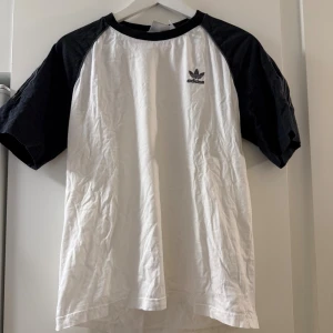 Adidas vit och svart t-shirt med logga - Snygg t-shirt från Adidas med vit framsida och svarta raglanärmar. Klassisk Adidas-logga broderad på bröstet och vita ränder på ärmarna. Rund hals och mjukt bomullsmaterial, perfekt för en sportig och chill stil.