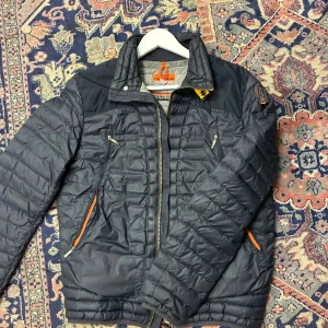 Mörkblå pufferjacka från Parajumpers - Säljer en mörkblå pufferjacka från Parajumpers i storlek M. Jackan har quiltad design, dragkedja framtill, flera fickor med dragkedja och en snygg patch på ärmen. Insidan är ljusgrå och detaljerna är orange och gul. Perfekt för kalla dagar.