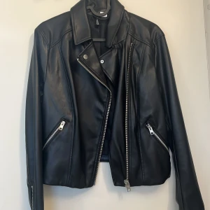 Svart skinnjacka från H&M med dragkedjor - Snygg svart skinnjacka från H&M med klassisk bikerstil. Jackan har silvriga dragkedjor, bred krage och två snedställda fickor med dragkedja. Perfekt för dig som vill ha en edgy look med stilrena detaljer.