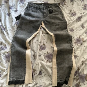 Amicci Maranello jeans grå, strl 32 - Grå jeans från Amicci i modellen Maranello med raka ben och beige paneler längs sidorna. Jeansen har diskreta slitningar, vita färgstänk och klassisk femficksdesign. Materialet är denim och passformen är rak och relaxed.
