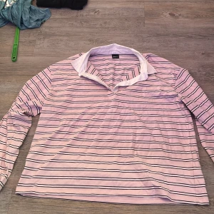 Randig pikétröja från Gina Tricot - Säljer en rosa och vit randig pikétröja från Gina Tricot i storlek L. Tröjan har klassisk vit krage, knappar framtill och långa ärmar. Perfekt för en chill och sportig look. Materialet är mjukt och känns skönt mot huden.