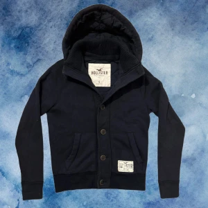 Vintage Hollister Kofta/Cardigan Med Knappar - Otroligt Vintage Svart hoodiejacka från Hollister med quiltat foder och stor huva. Jackan har ribbad krage, knappstängning (sista knappen är halvt sönder, fungerar fortfarande jätte bra och syns knappast då den ligger längst ner) och två fickor fram. Märkespatch på bröstet och insidan. Denna är riktigt vintage och riktigt svår att finna! Längd 63cm, Bredd under armhåla 50cm! Modell 175, 70kg Pris kan diskuteras vid snabbt köp!