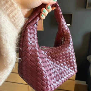 Vinröd flätad handväska i skinn - Säljer en vinröd handväska i flätat skinn med mjuk, rundad form och kort handtag. Väskan har dragkedja upptill och en stilren look som sticker ut. Perfekt storlek för det viktigaste och ger en cool touch till din outfit.
