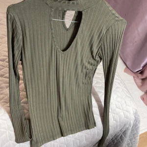 Olivgrön ribbad topp från Atmosphere - Säljer en olivgrön ribbad topp från Atmosphere med lång ärm och snygg cut out-detalj vid halsen. Toppen har hög krage med två knappar bak och är tillverkad i mjukt, stretchigt material. Perfekt för att lyfta din outfit med en cool touch.