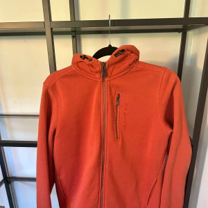 Orange hoodie från Sail Racing - Säljer en orange hoodie från Sail Racing med dragkedja framtill och bröstficka med zip. Tröjan har huva med snörning och långa ärmar. Perfekt för dig som gillar sportig och avslappnad stil.