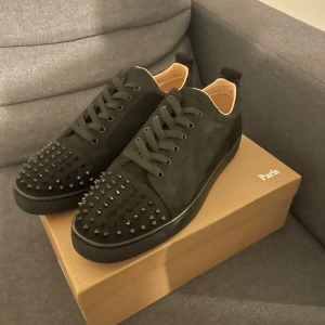 Christian Louboutin svarta sneakers med nitar - Svarta sneakers från Christian Louboutin med coola svarta nitar på tån och klassisk röd sula. Skorna har snörning och är tillverkade i mocka med skinnfoder. Snygg, lyxig design som sticker ut och passar perfekt för dig som gillar en stilren look.