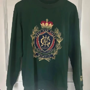 Unika Gant Sweatshirt  - Mörkgrön sweatshirt från Gant med stort broderat emblem i guld, rött och blått på bröstet och mindre brodyr på ärmen. Tröjan har relaxed fit, rund hals och långa ärmar. Perfekt för dig som gillar preppy och klassisk stil.