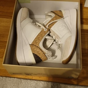 Michael Kors Georgie Trainer sneakers - Snygga Michael Kors Georgie Trainer sneakers i vitt och beige med MK-logga på sidan och tungan. Skorna har en mix av skinn, mocka och textil, samt bruna detaljer med MK-mönster. Platt gummisula och snörning framtill. Perfekt för dig som gillar stilrena och trendiga sneakers.