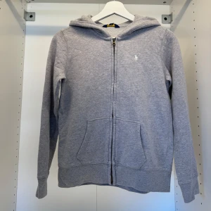 Grå hoodie från Polo Ralph Lauren - Grå hoodie från Polo Ralph Lauren med dragkedja framtill, huva och känguruficka. Liten vit broderad logga på bröstet. Skön och enkel stil som funkar året runt. Perfekt för chill dagar eller när du vill ha något bekvämt.