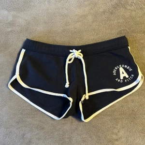 Mörkblåa mini shorts Abercrombie & Fitch XS - Snygga mörkblåa shorts från Abercrombie & Fitch i storlek XS. De har vit snörning i midjan, vita kanter och en broderad logga på ena benet. Mjukt bomullsmaterial och sportig passform med rundade benkanter. Perfekta för chill eller träning.
