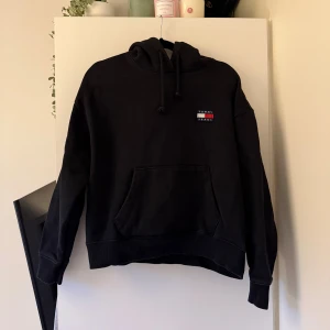 Svart hoodie från Tommy Jeans - Svart hoodie från Tommy Jeans med klassisk logga på bröstet. Tröjan har huva med snörning och en stor magficka framtill. 