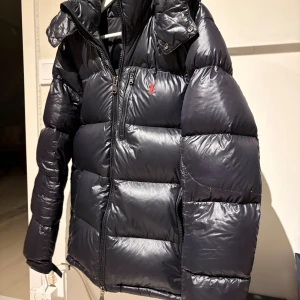  Ralph Lauren jacka - Säljer en svart pufferjacka från Polo Ralph Lauren med huva och dragkedja framtill. Jackan har en stilren design med diskret logga på bröstet och breda, quiltade sektioner. Perfekt för kalla dagar och har en riktigt schysst look.