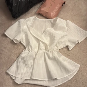 Vit omlottblus med volang - Säljer en vit blus med omlottdesign och volangkant. Blusen har korta ärmar och ett lätt rynkat midjeparti som ger en snygg siluett. Materialet är lätt och har en subtil ribbad struktur som ger extra känsla. Står inte vart den är köpt och jag minns inte vart💕💕 