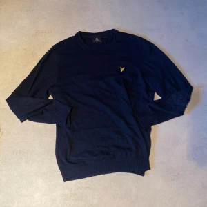 Stickad tröja från Lyle&Scott - Snygg marinblå tröja från Lyle & Scott med rund halsringning och gul logga broderad på bröstet. Tröjan har långa ärmar och ribbade muddar vid ärmslut och nederkant. Perfekt för en stilren och klassisk look. 