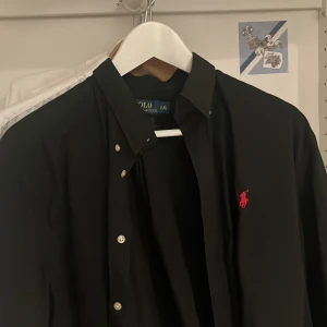 Svart skjorta från Polo Ralph Lauren - Svart skjorta från Polo Ralph Lauren med klassisk krage och knappar framtill. Röd broderad logga på bröstet ger en snygg kontrast. Skjortan är långärmad och har en stilren, tidlös design som passar till många olika looks.