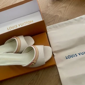 Louis Vuitton vita sandaler - Snygga vita sandaler från Louis Vuitton med låg klack och öppen tå. Skorna har en beige detalj med guldfärgad knapp och diskret rosa söm. Materialet är skinn med textilstruktur och logotypen är präglad på innersulan. Perfekt för dig som vill ha en lyxig och stilren look❤️