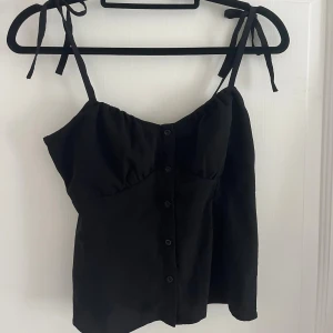 Svart linnetopp med knyt från Bubbleroom - Svart linnetopp från Bubbleroom med knytbara axelband, smockad rygg och knappar framtill. 