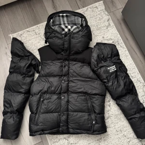 Svart pufferjacka från Burberry - Säljer en helt ny svart pufferjacka från Burberry med avtagbara ärmar, jackan kan alltså bli till en väst. Jackan har ett stort Burberry London England-märke på ärmen och rutigt foder i huvan. Två dragkedjefickor framtill och quiltad design. Perfekt för kalla dagar. För mer funderingar/bilder skriv privat 