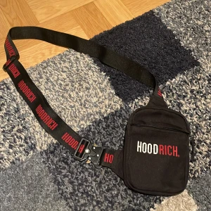 Svart axelväska från Hoodrich - Snygg svart axelväska från Hoodrich med justerbart band där loggan är tryckt i rött och vitt. Väskan har en dragkedja framtill och är perfekt för att bära dina viktigaste grejer. Stilren och street-inspirerad design.