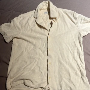 Beige kortärmad skjorta från Zara - Snygg beige skjorta från Zara i storlek S. Skjortan har korta ärmar, klassisk krage och knäppning framtill med matchande knappar. Materialet är mjukt och har en frottéliknande struktur som ger en chill vibe. Perfekt för varma dagar när du vill vara både bekväm och stilren.