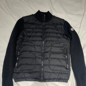 Moncler Cardigan Jacka - Svart Cardigan jacka från Moncler i storlek S. Jackan har quiltad front i dun och släta, stickade ärmar med Moncler-logga på vänster arm. Två dragkedjefickor framtill och hel dragkedja. Perfekt för hösten när du vill ha både stil och värme.