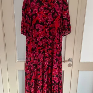 Blommig långklänning från Zizzi - Säljer en färgstark långklänning från Zizzi med blommigt mönster i rött, rosa och svart. Klänningen har korta ärmar och ett flowy fall som ger en chill vibe. Perfekt för dig som gillar att sticka ut med coola färger och mönster.