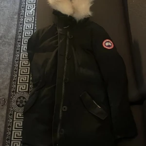 Svart dunjacka från Canada Goose - Säljer en svart dunjacka från Canada Goose med klassisk patch på ärmen och snygg pälskantad huva. Jackan har dragkedja och knappar framtill samt två snedställda fickor. Perfekt för kalla vinterdagar och riktigt varm.