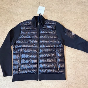 Svart Cardigan Moncler  - Snygg svart Cardigan från Moncler med glansig quiltad framsida och ärmar i stickad bomull. Jackan har dragkedja framtill, två fickor med dragkedja och en Moncler-logga på ärmen. Perfekt för dig som vill ha en stilren och exklusiv look.
