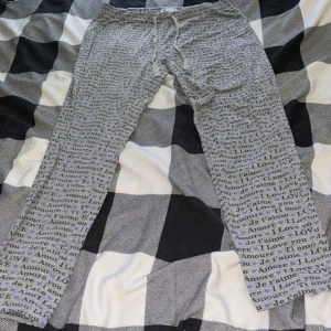 Grå mjukisbyxor med textmönster - Grå mjukisbyxor från C i storlek M med snörning i midjan. Byxorna har ett all-over mönster med ord som 'LOVE', 'Je t'aime', 'Ti amo' och små lila hjärtan. Perfekta för chill dagar hemma eller när du vill ha något bekvämt.