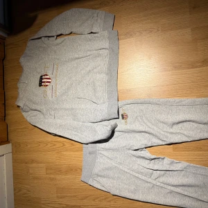 Grå GANT hoodie med broderad logga - Snygg ljusgrå hoodie från GANT med broderad logga och text på bröstet i guld och rött. Tröjan har känguruficka framtill och ribbade muddar vid ärmslut och nederkant. Perfekt för chill dagar eller när du vill ha en sportig look.