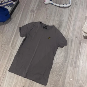 Grå t-shirt från Lyle & Scott - Säljer en stilren grå t-shirt från Lyle & Scott med klassisk gul logga på bröstet. T-shirten är kortärmad och har en normal passform. Perfekt till jeans eller shorts för en clean look.