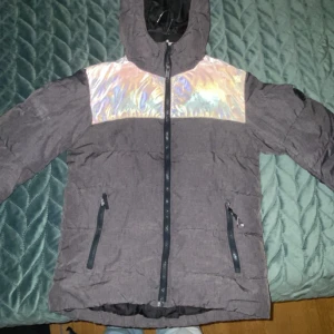 Grå pufferjacka med holografisk detalj - Säljer en grå pufferjacka från Jack & Jones med en riktigt fet holografisk panel över bröstet. Jackan har huva, hel dragkedja framtill och två snedställda fickor med dragkedja. Perfekt för dig som vill sticka ut i vinterkylan.