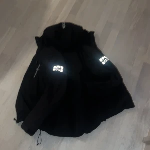 Svart Sail Racing dunjacka med reflex - Säljer en svart dunjacka från Sail Racing med reflekterande loggor på ärmarna. Jackan har dragkedja framtill, justerbar huva och quiltad insida. Perfekt för dig som vill ha en stilren och funktionell jacka med snygga detaljer.