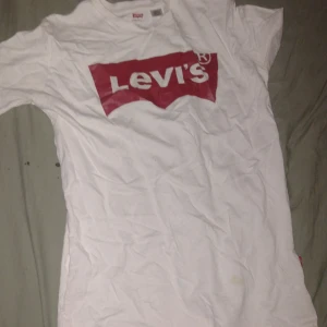 Vit Levi's t-shirt med röd logga - Vit t-shirt från Levi's i storlek S med den klassiska röda loggan tryckt framtill. T-shirten är kortärmad och har en rund halsringning. Tillverkad i mjuk bomull för en skön känsla. Perfekt för en avslappnad och stilren look.