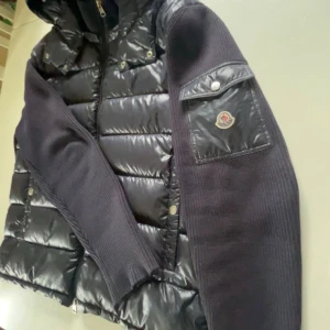 Svart cardigan från Moncler med huva - Snygg svart dunväst från Moncler med glansig quiltad framsida och stickade ärmar. Västen har huva, dragkedja och en ikonisk Moncler-logga på ärmen. Materialmix av dun, polyester och stickat ger en modern look. Perfekt för lager-på-lager-stil.