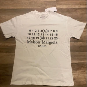 Maison Margiela vit t-shirt med tryck - Vit t-shirt från Maison Margiela med ikoniskt siffertryck och logga på bröstet. Klassisk passform, rund hals och korta ärmar. Tillverkad i mjuk bomull för en skön känsla. Perfekt statement-plagg för dig som gillar stilrena designer.
