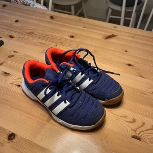 Blå Adidas sneakers med vita ränder - Säljer ett par blå Adidas sneakers med klassiska vita ränder på sidorna och orange detaljer på insidan. Skorna har snörning, gummisula i brunt och inslag av vitt och orange undertill. Perfekta för inomhussport. (Tror att modellen är ”adiwear” eller nått😅)